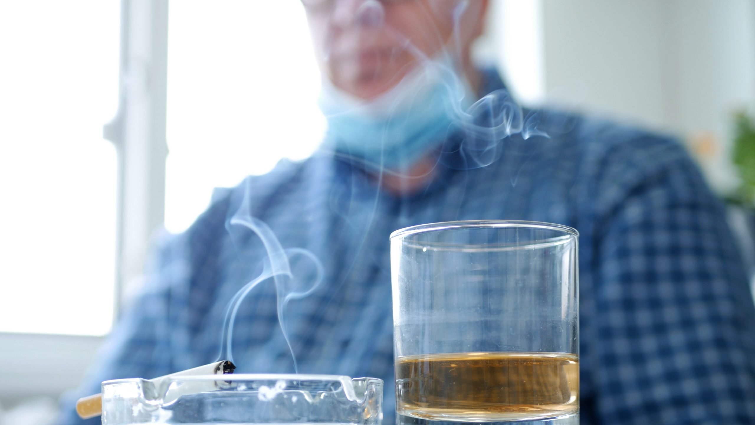 Man in COVID-19 mask drinks and smokes alone.