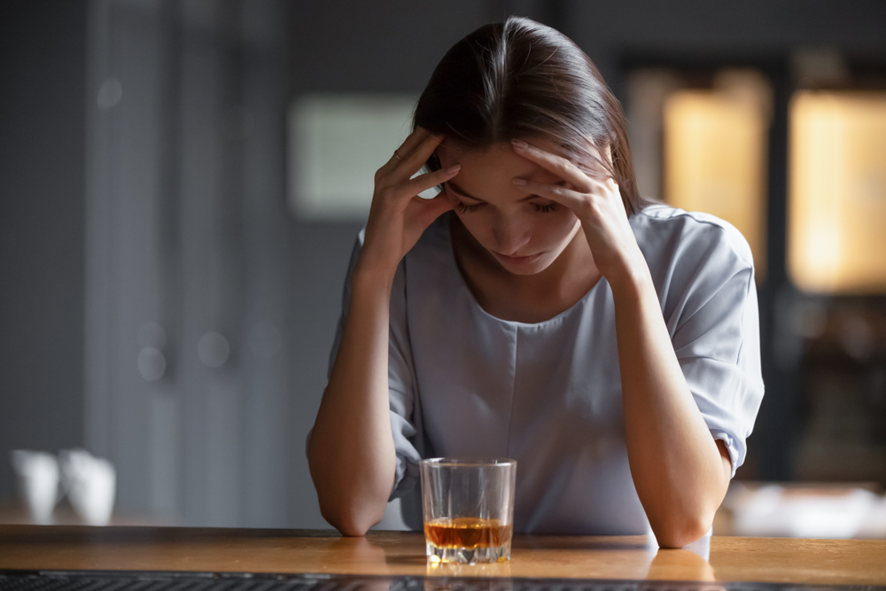 Woman has head in hands next to glass of whisky.